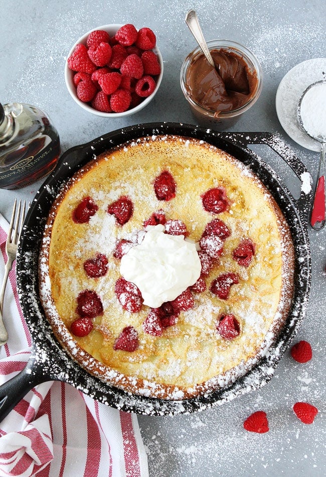 Raspberry Dutch Baby Recipe on twopeasandtheirpod.com This puffy oven baked pancake is dotted with fresh raspberries, dusted with powdered sugar, and topped with whipped cream. It is a real breakfast treat! Dutch Baby Pancake in skillet with ingredients and toppings