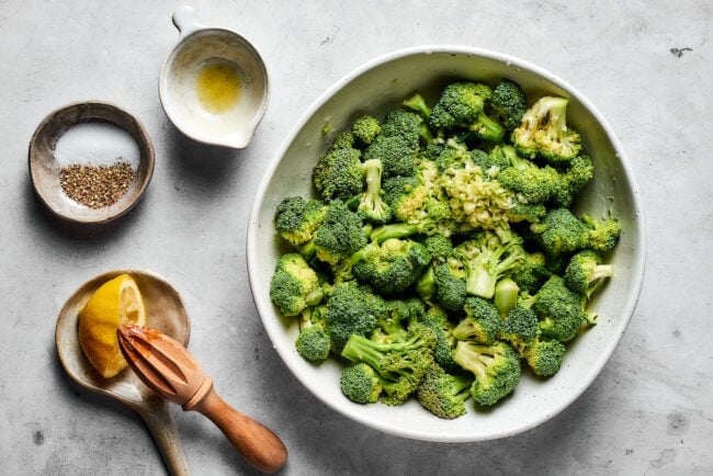 Easy roasted broccoli with garlic is destined to be your new go-to side dish! This crispy, oven-baked broccoli recipe is even kid-approved. A bowl of broccoli florets next to a bowls with juiced lemon, seasoning, and garlic.