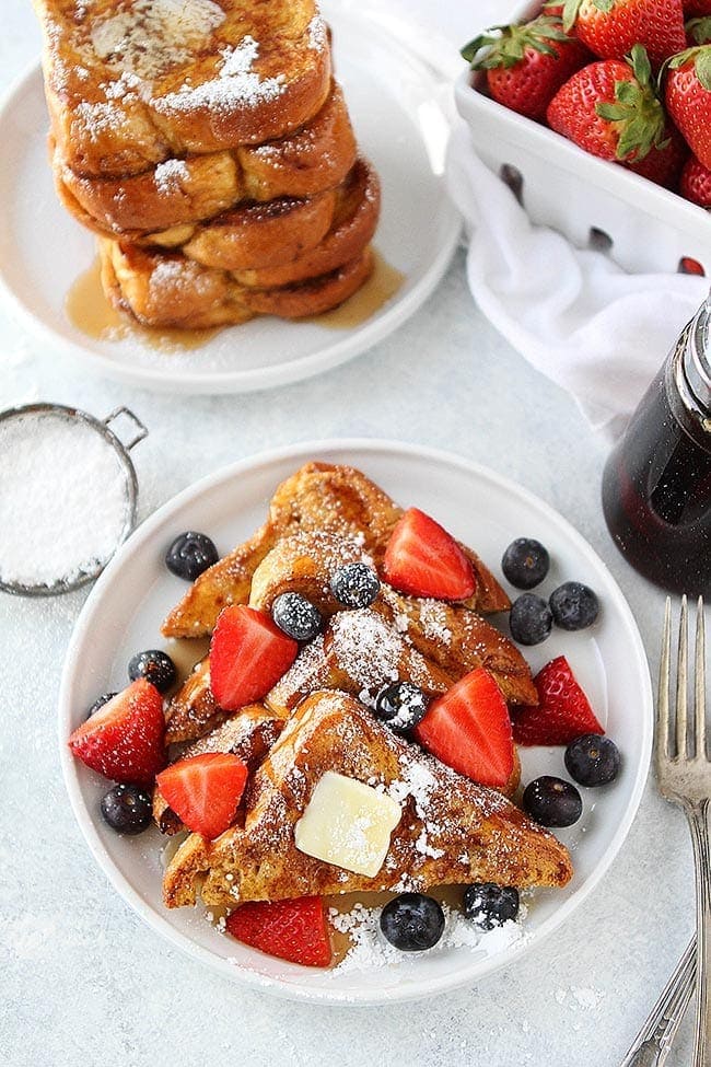 The BEST French Toast recipe and it's easy to make at home! #frenchtoast #breakfast #brunch #easyrecipe #bread #familyfavorite #vegetarian French Toast cut in slices on plate with powdered sugar, butter, syrup, and berries.