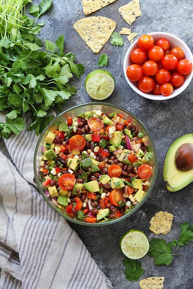 Easy Black Bean Salad with corn, avocado, tomatoes, red pepper, cilantro, and lime. The perfect summer side dish to any meal! You can also serve it as a dip with tortilla chips! #blackbean #vegetarian #vegan #glutenfree #easyrecipe #healthyrecipe #avocado #corn #summer #saladrecipe Black bean salad in bowl with cilantro, tomatoes, lime, avocado, and chips.