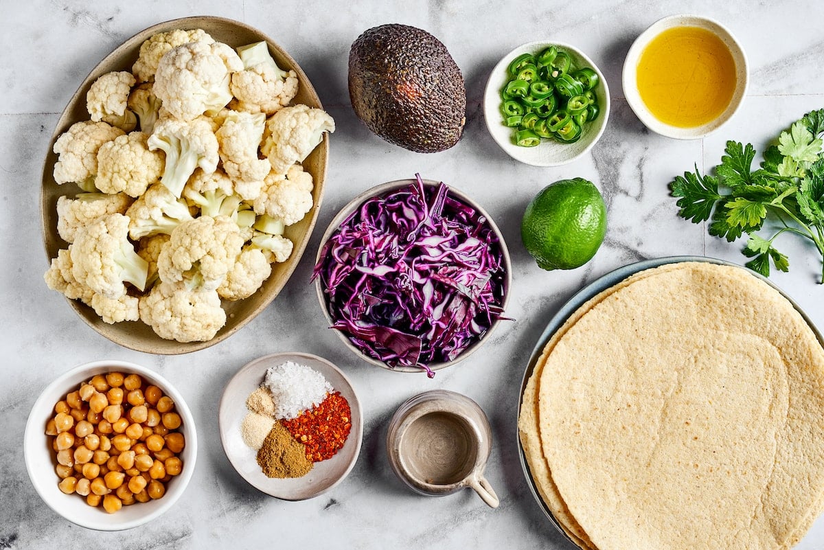 These roasted cauliflower and chickpea tacos are spicy, fresh, bursting with flavor, and a healthy vegetarian Mexican dinner Overhead view of ingredients for roasted cauliflower and chickpea tacos: tortillas, raw cauliflower, car cabbage, jalapeño slices, a lime, an avocado, canned chicpeas, a bowl of spices, a jar of water, a bowl of olive oil, and a sprig of cilantro
