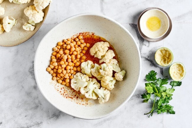 These roasted cauliflower and chickpea tacos are spicy, fresh, bursting with flavor, and a healthy vegetarian Mexican dinner A bowl of marinade with chickpeas and raw cauliflower on top, next to a bowl of cauliflower, squeezed lime halves, cilantro, and a stack of empty bowls