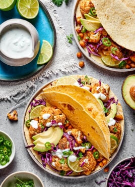 A plate with two roasted cauliflower and chickpea tacos on it, topped with avocado and fresh jalapeรฑo, next to a plate with lime slices and a jar of lime crema, with half an avocado and a plate of tacos next to it
