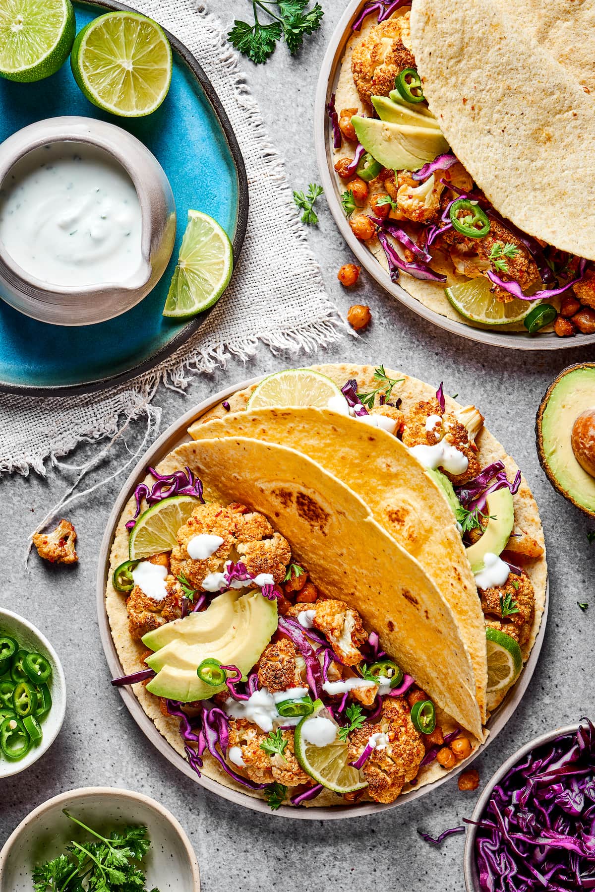 These roasted cauliflower and chickpea tacos are spicy, fresh, bursting with flavor, and a healthy vegetarian Mexican dinner A plate with two roasted cauliflower and chickpea tacos on it, topped with avocado and fresh jalapeño, next to a plate with lime slices and a jar of lime crema, with half an avocado and a plate of tacos next to it