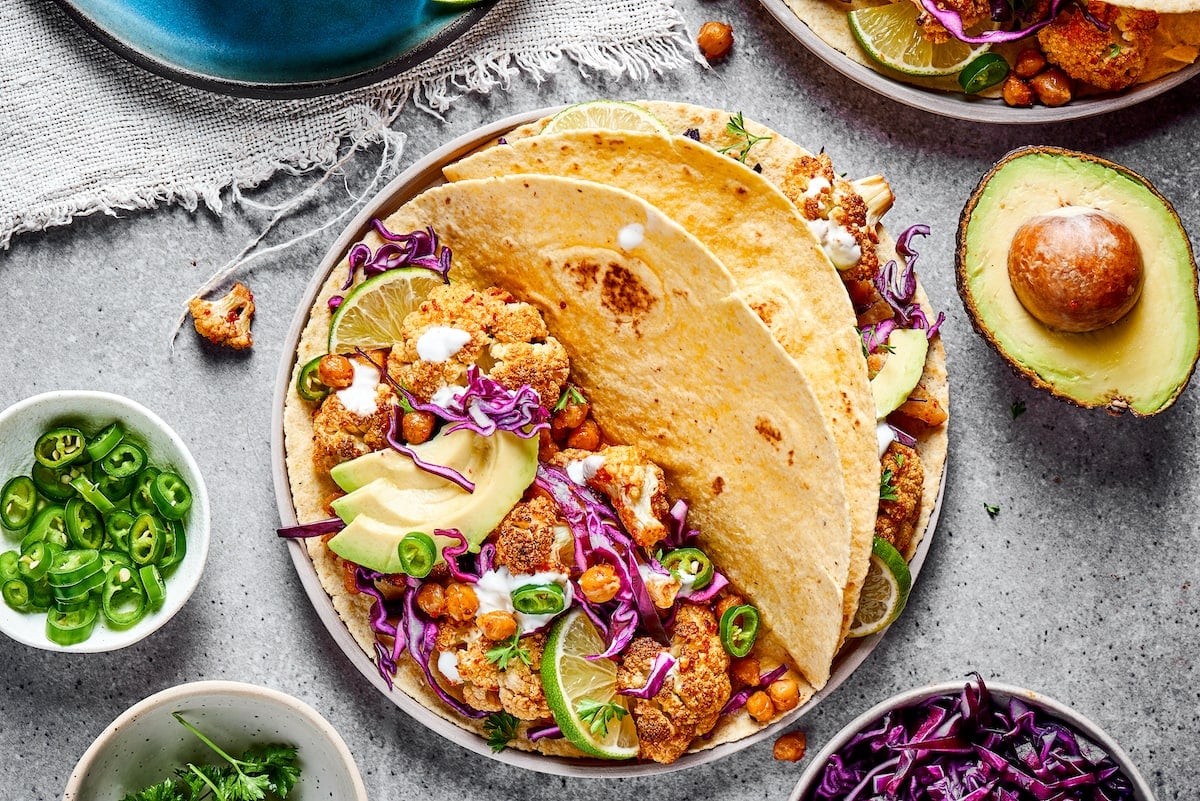 These roasted cauliflower and chickpea tacos are spicy, fresh, bursting with flavor, and a healthy vegetarian Mexican dinner A plate with two roasted cauliflower and chickpea tacos, next to half an avocado and a bowl of jalapeño slices