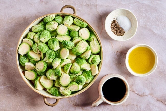 This roasted brussels sprouts recipe uses balsamic vinegar for an easy and flavorful Italian side dish that's healthy and vegetarian. A mixing bowl full of raw Brussels sprouts, next to a small container of balsamic vinegar, a small bowl of olive oil, and a small bowl of salt and pepper