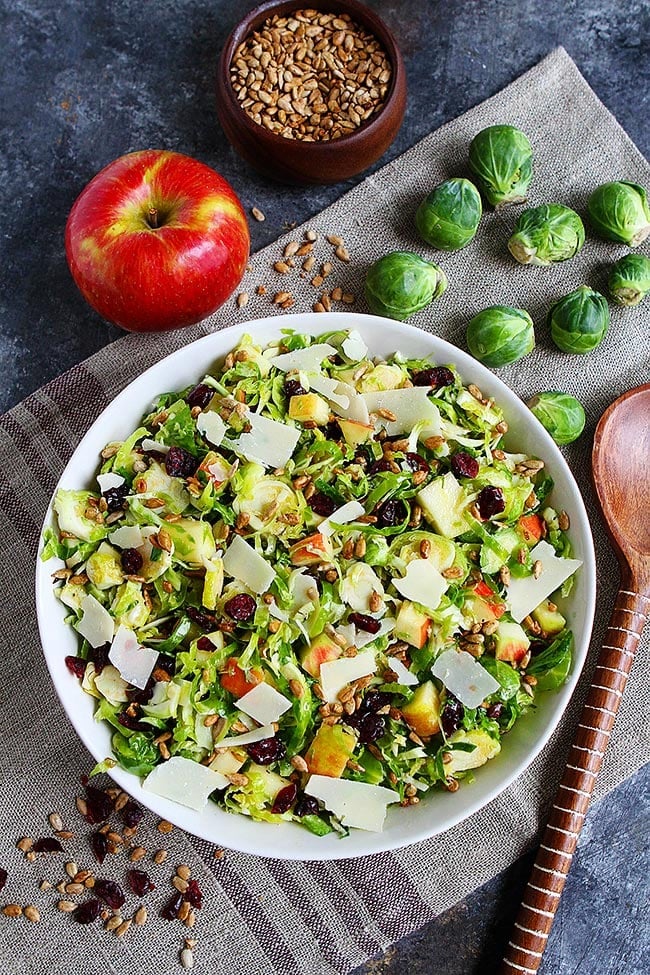 This raw shaved Brussels sprouts salad is crisp and refreshing, with apples and a maple dressing. It's perfect for Thanksgiving or Christmas! A bowl of shaved Brussels sprouts salad topped with shaved parmesan, on top of a place mat, next to a whole apple, nine whole Brussels sprouts, a wooden serving spoon, a bowl of sunflower seeds, and scattered sunflower seeds and dried cranberries.