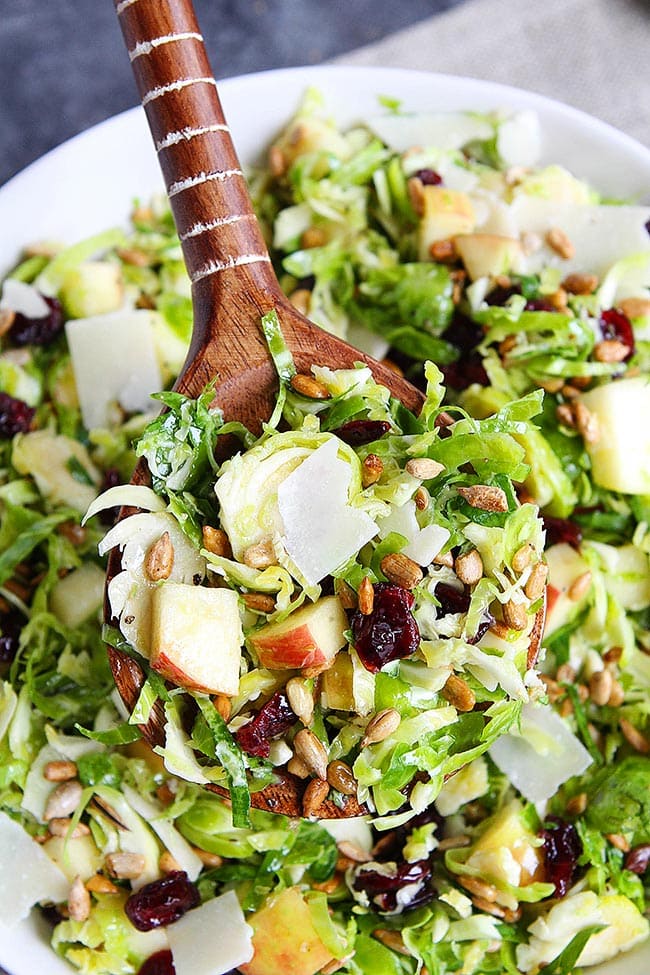 This raw shaved Brussels sprouts salad is crisp and refreshing, with apples and a maple dressing. It's perfect for Thanksgiving or Christmas! Overhead view of a wooden serving spoon full of shaved Brussels sprouts salad, with slices of apples, dried cranberries, sunflower seeds, and shaved parmesan cheese, with a bowl of the salad in the background.