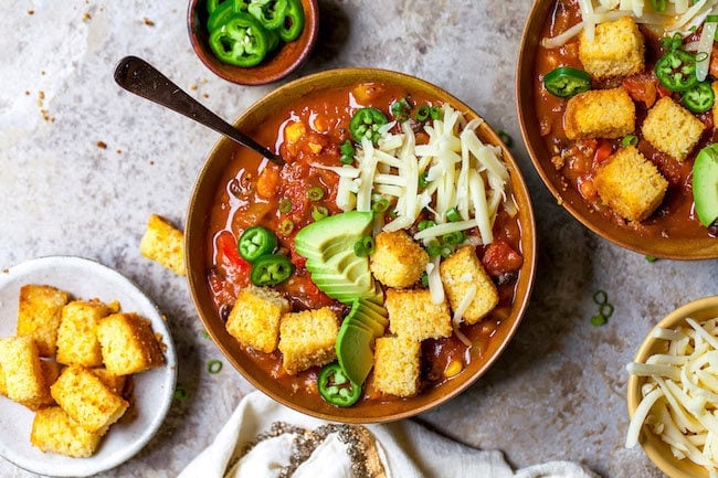 pumpkin chili in bowl with cornbread croutons, avocado, jalapeño slices, and cheese.