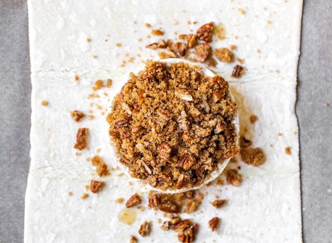 wheel of brie cheese on top of a sheet of puff pastry with pecans and brown sugar on top.