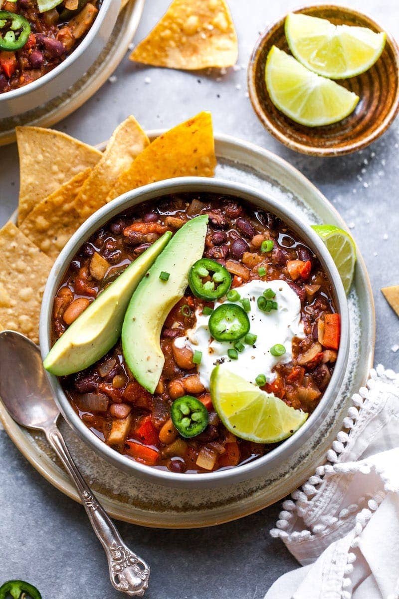 Vegetarian Chili in bowl with avocado slices, sour cream, jalapeño slices, and lime wedge and tortilla chips on the side.