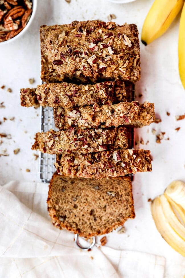 banana nut bread cut into slices on cooling rack with tea towel and bananas and nuts in bowl.