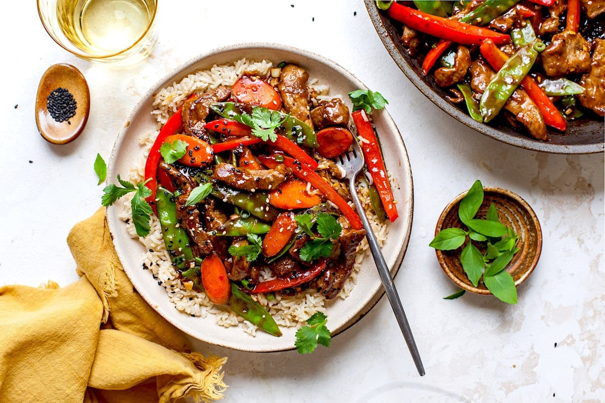 beef stir fry in bowl with vegetables, rice, and fork and stir fry in skillet.
