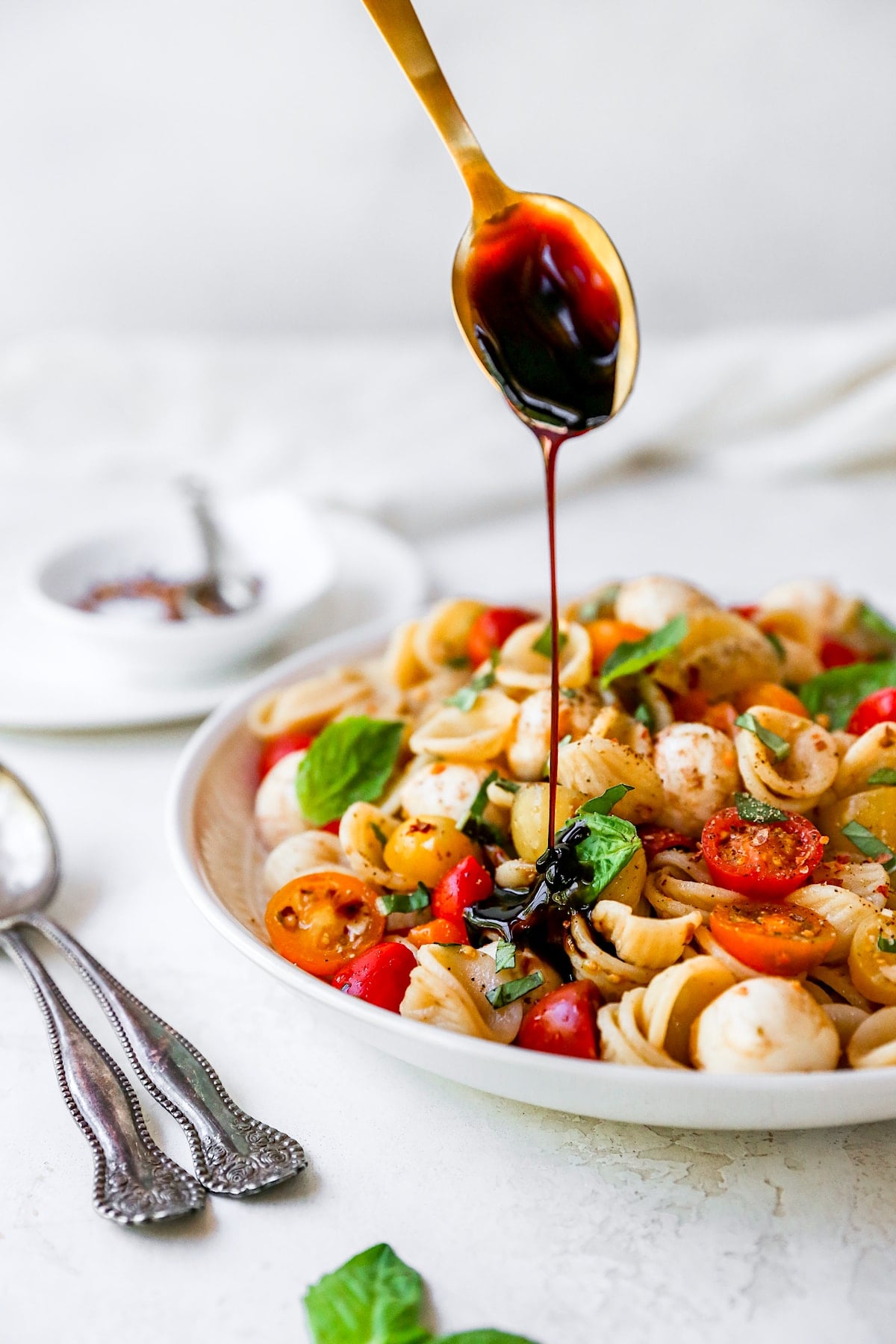 caprese pasta salad in bowl with balsamic glaze being drizzled over the top with spoon.
