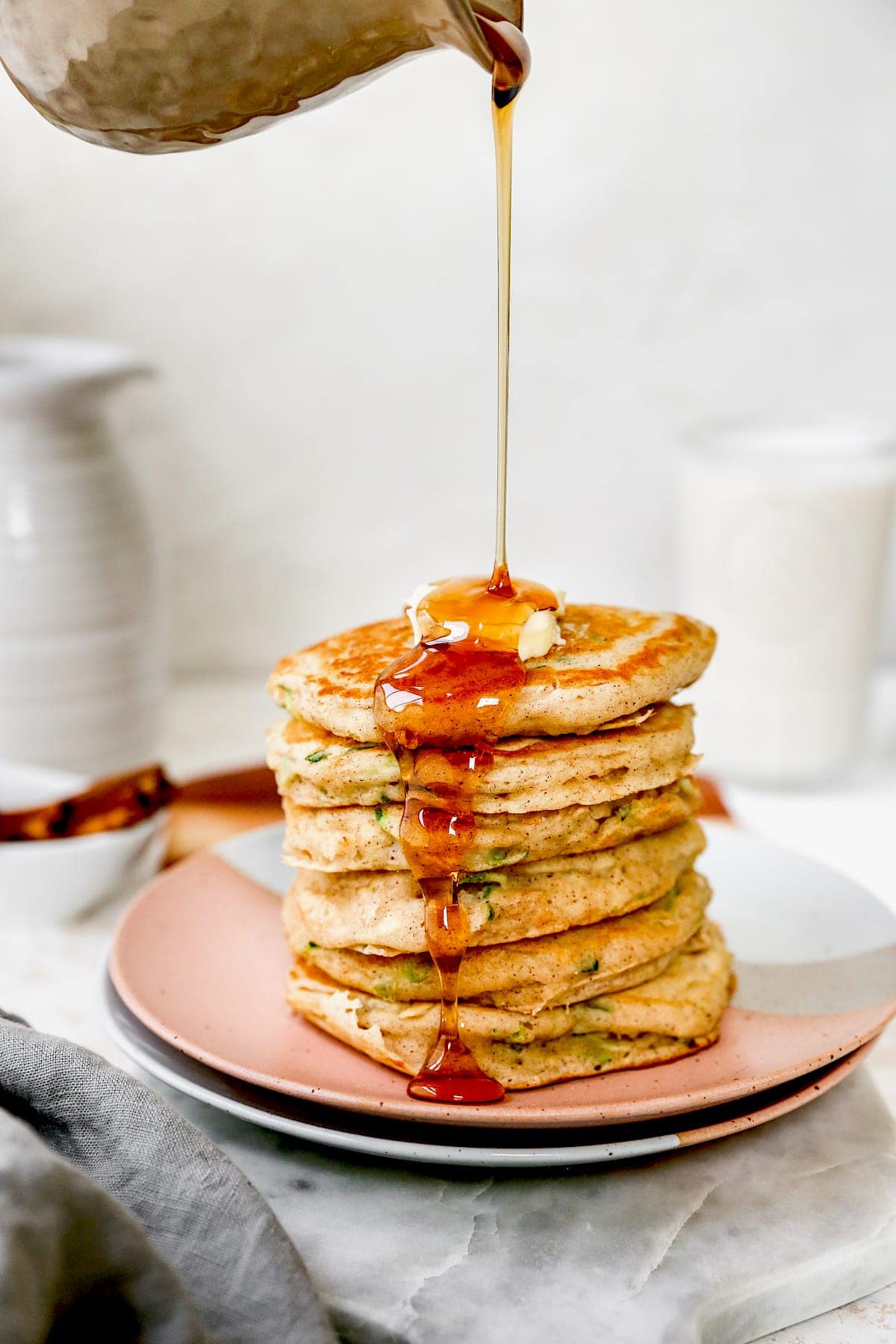 stack of zucchini pancakes on plate with butter and maple syrup being poured on top.