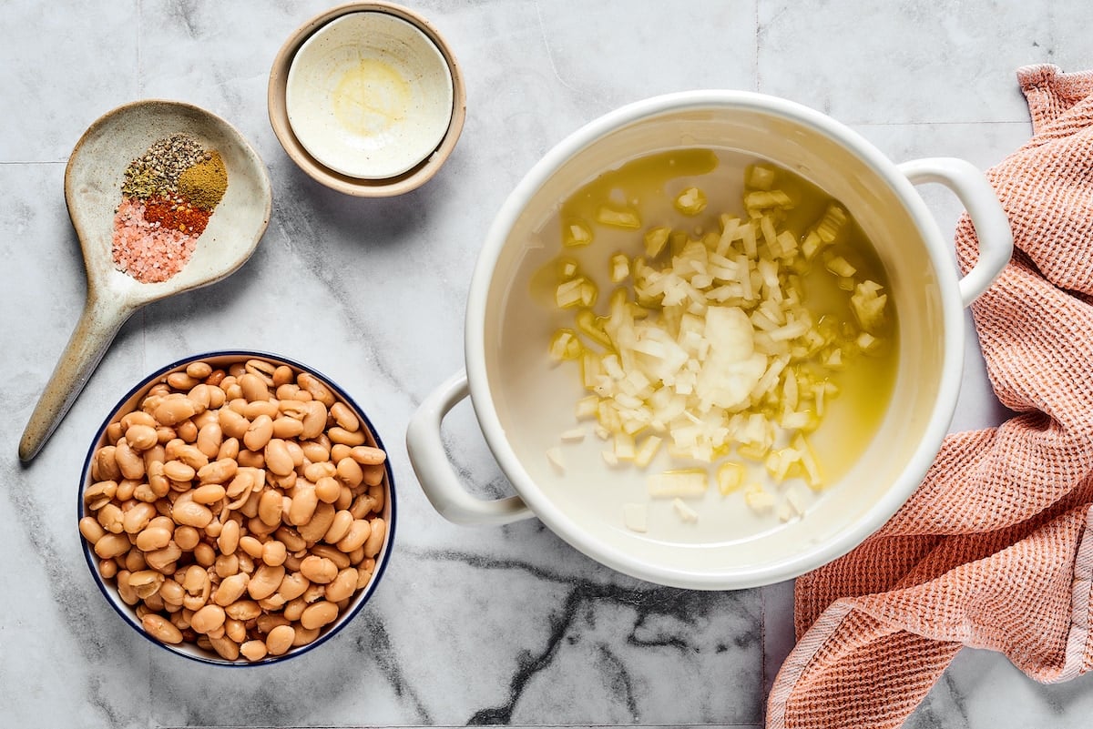 This comforting white chicken chili is easy to make with white beans, zesty seasonings, and rotisserie chicken. A favorite weeknight meal! Sautéed garlic and onions in a large pot next to bowls of chili ingredients.