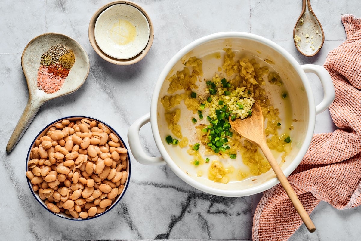 This comforting white chicken chili is easy to make with white beans, zesty seasonings, and rotisserie chicken. A favorite weeknight meal! Sautéed garlic, onions, and jalapeños in a large pot next to bowls of chili ingredients.