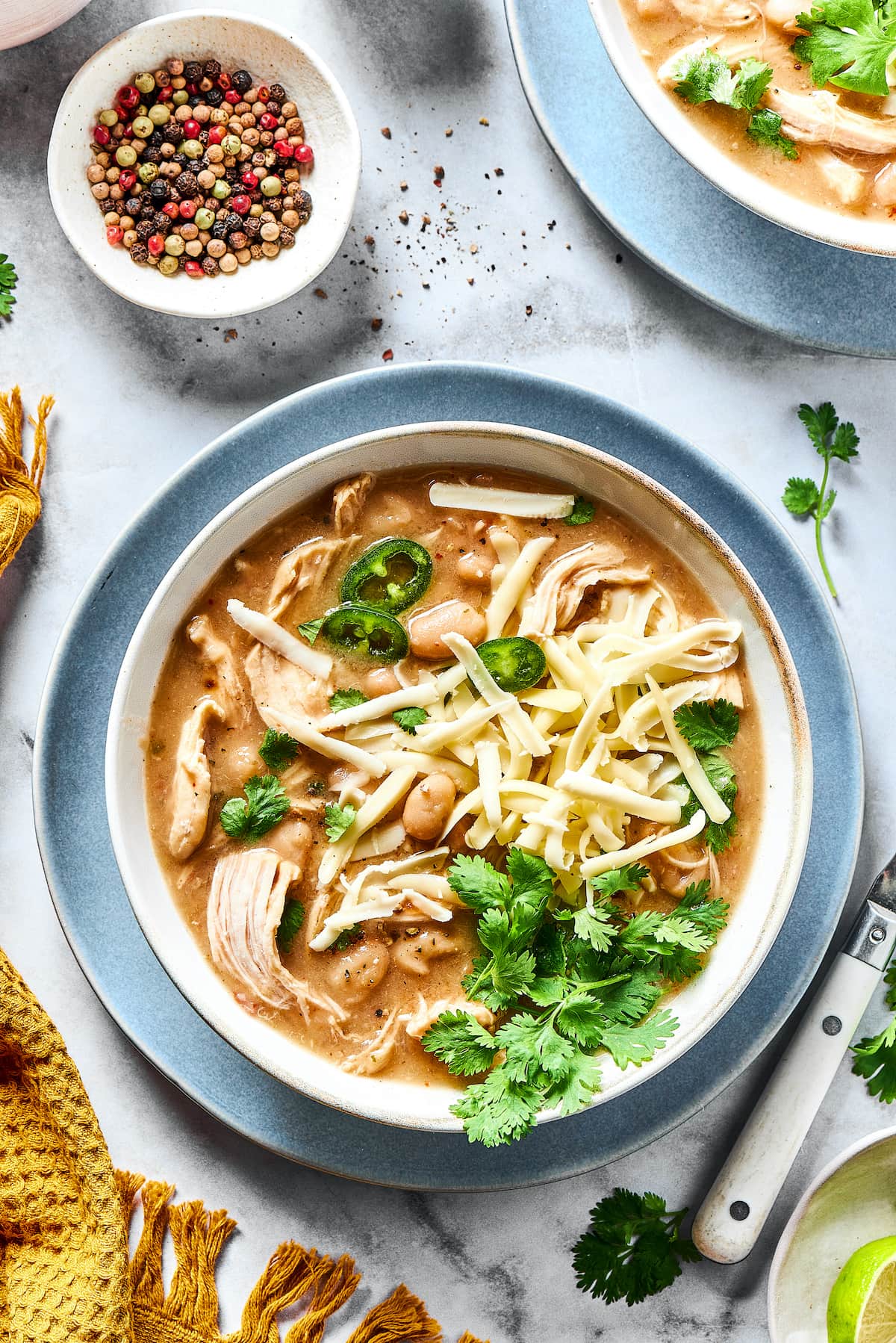 This comforting white chicken chili is easy to make with white beans, zesty seasonings, and rotisserie chicken. A favorite weeknight meal! Overhead view of a bowl of white chicken chili garnished with fresh cilantro, next to a small bowl of rainbow peppercorns.