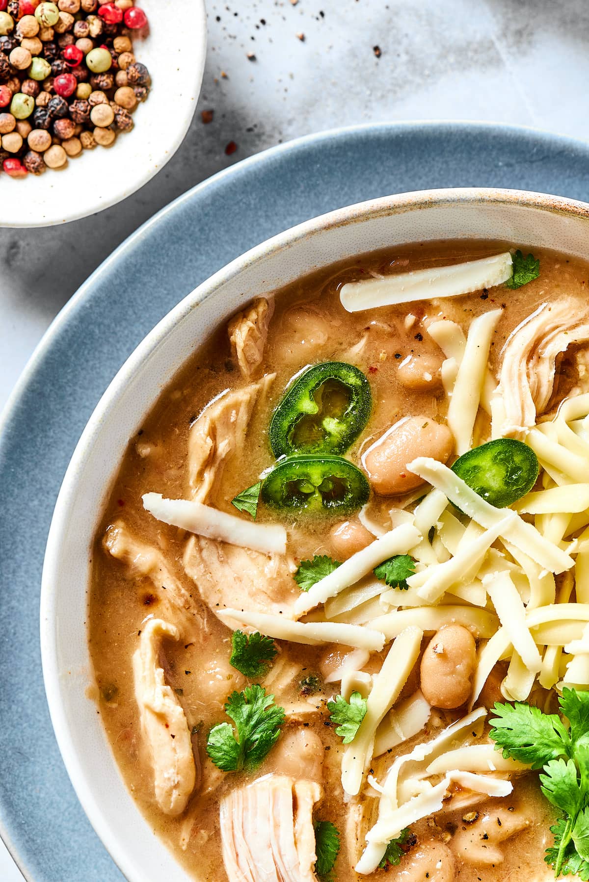 This comforting white chicken chili is easy to make with white beans, zesty seasonings, and rotisserie chicken. A favorite weeknight meal! Close up overhead view of a bowl of white chicken chili garnished with fresh cilantro, next to a small bowl of rainbow peppercorns.