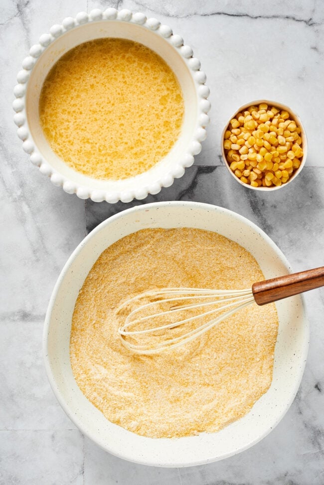 This easy cornbread recipe is tender, moist, perfectly sweet, and takes only 35 minutes to make, making it a wonderful side dish A bowl with cornmeal, flour, salt, sugar, baking soda, and baking powder, mixed together with a whisk in the bowl, next to a bowl with eggs, buttermilk, melted butter, and oil, mixed together, next to a bowl of corn