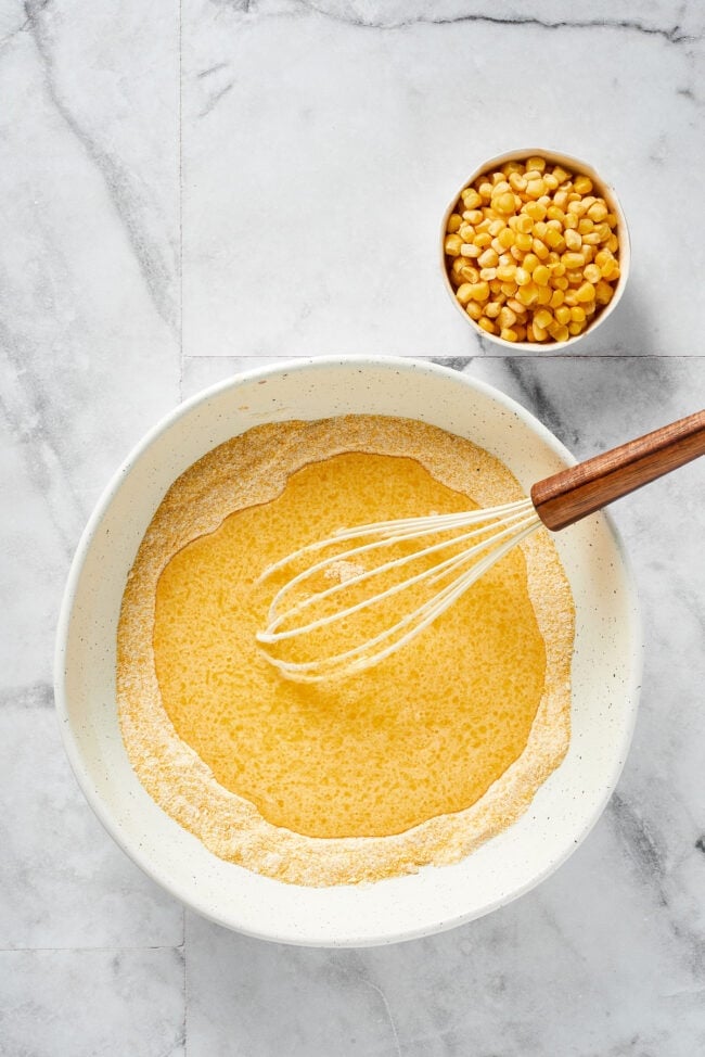 This easy cornbread recipe is tender, moist, perfectly sweet, and takes only 35 minutes to make, making it a wonderful side dish A bowl of the dry ingredients for cornbread, with the wet ingredients poured on top, with a whisk in the bowl, next to a bowl of corn