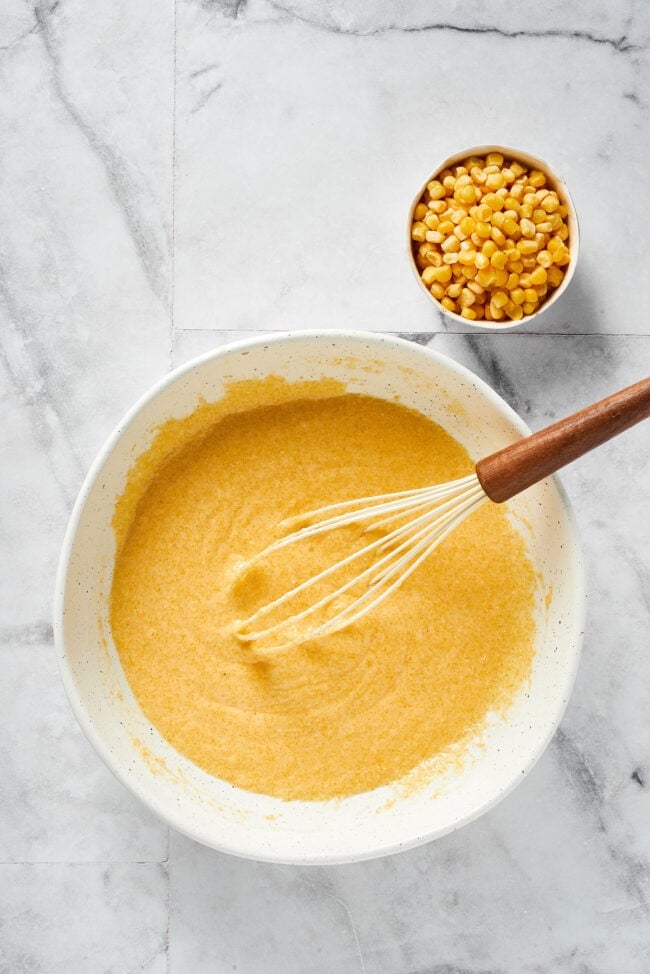 This easy cornbread recipe is tender, moist, perfectly sweet, and takes only 35 minutes to make, making it a wonderful side dish A mixing bowl filled with cornbread batter, without the corn, with a whisk in it, next to a bowl of corn kernels