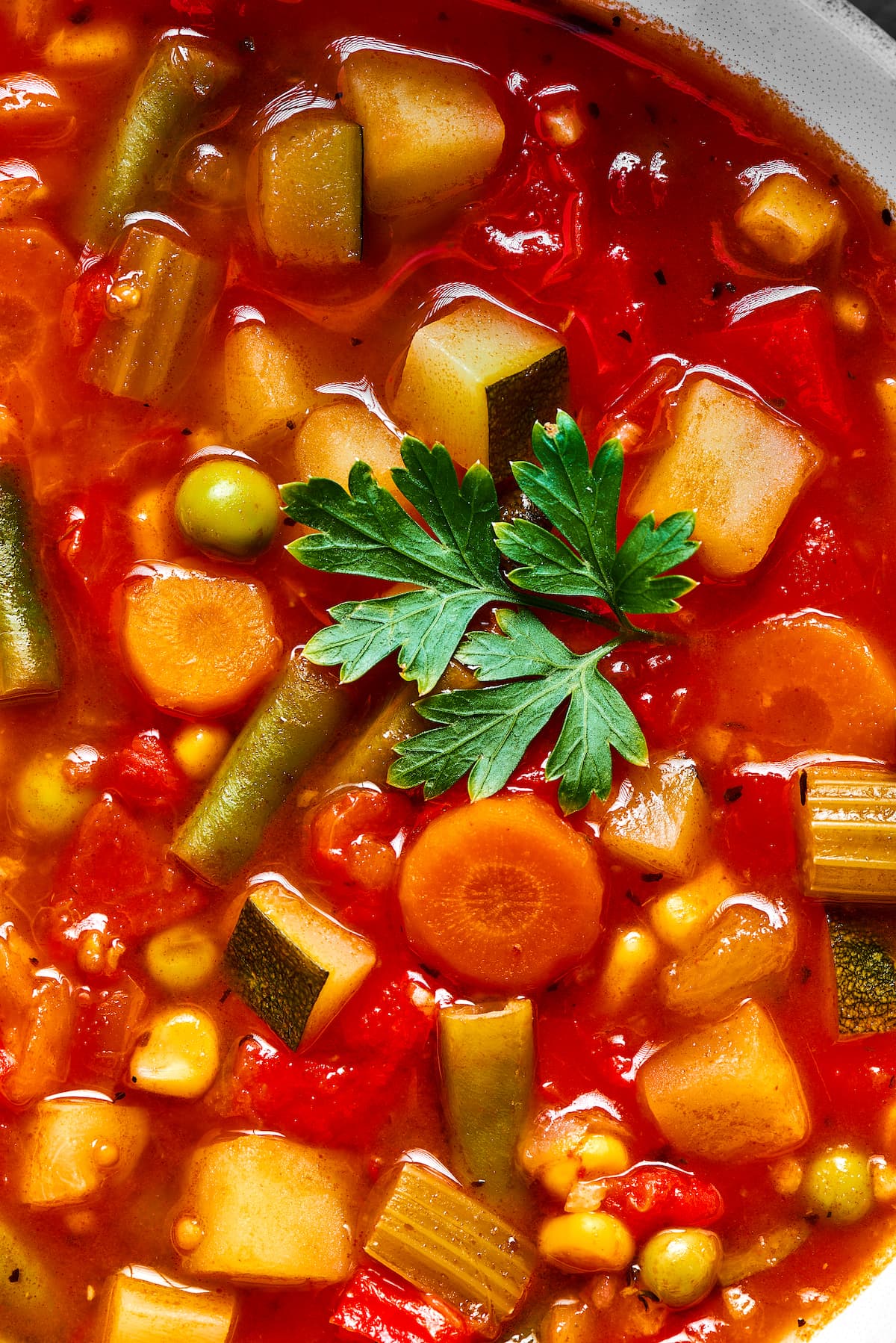 A bowl of homemade vegetable soup with a spoon, next to a large pot. Close up of homemade vegetable soup garnished with a green parsley leaf.