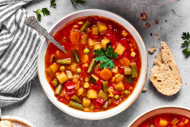 This comforting homemade vegetable soup recipe is a family favorite that's so much better than canned soup! Enjoy a bowl for lunch or dinner. A bowl of homemade vegetable soup with a spoon, next to a bread slice.