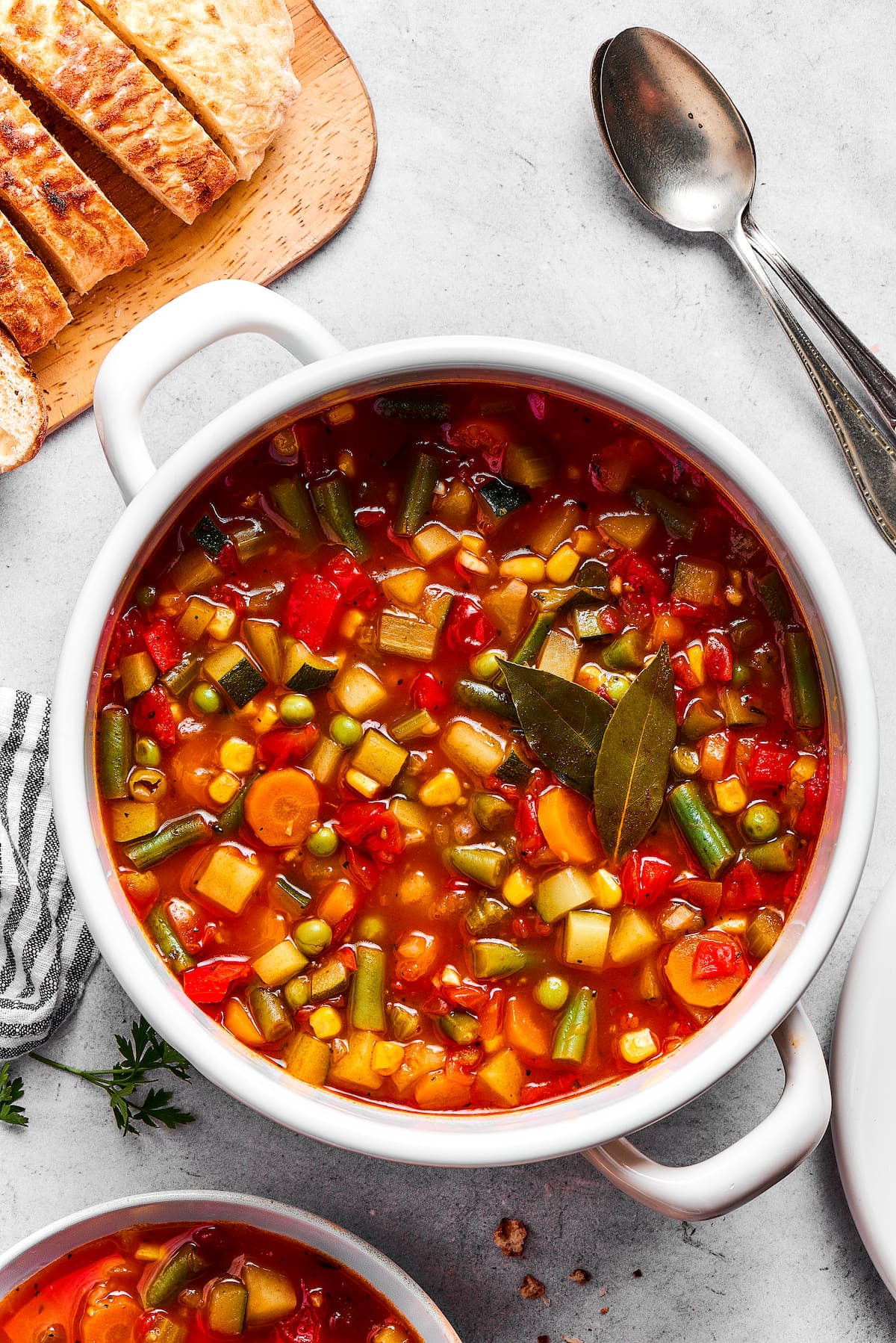 A bowl of homemade vegetable soup with a spoon, next to a large pot. A pot of vegetable soup next to a loaf of crusty bread and a spoon.