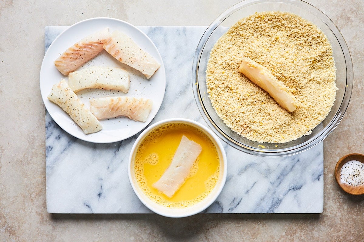 white fish sticks on plate, white fish in egg mixture, white fish stick in panko mixture.