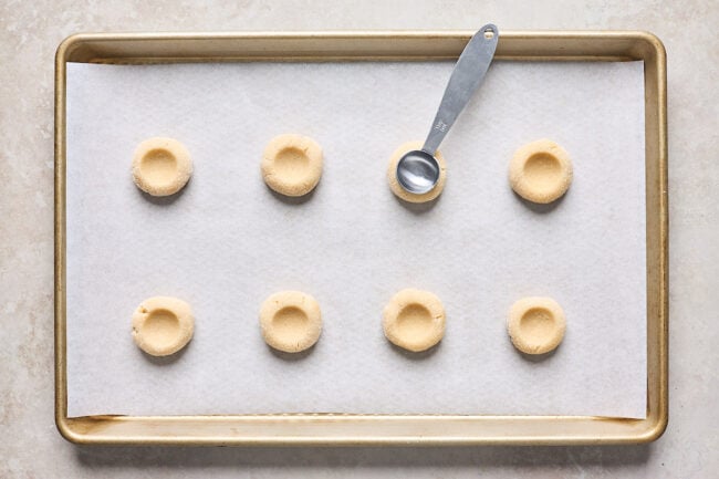 thumbprint cookie dough pressed into thumbprints with a teaspoon on a baking sheet lined with parchment paper.