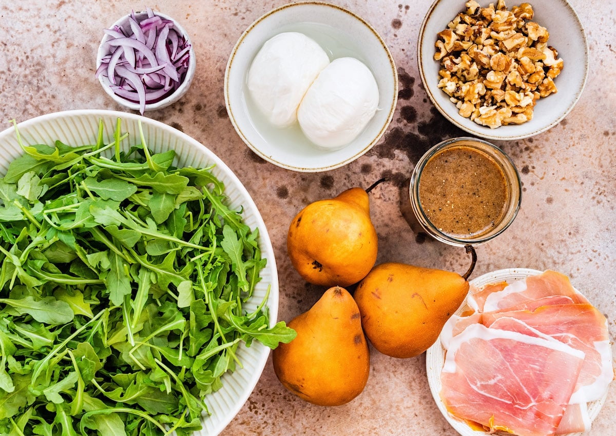 arugula in a bowl, pears, prosciutto on plate, balsamic vinaigrette in a jar, burrata cheese, red onion, and chopped walnuts in a bowl.