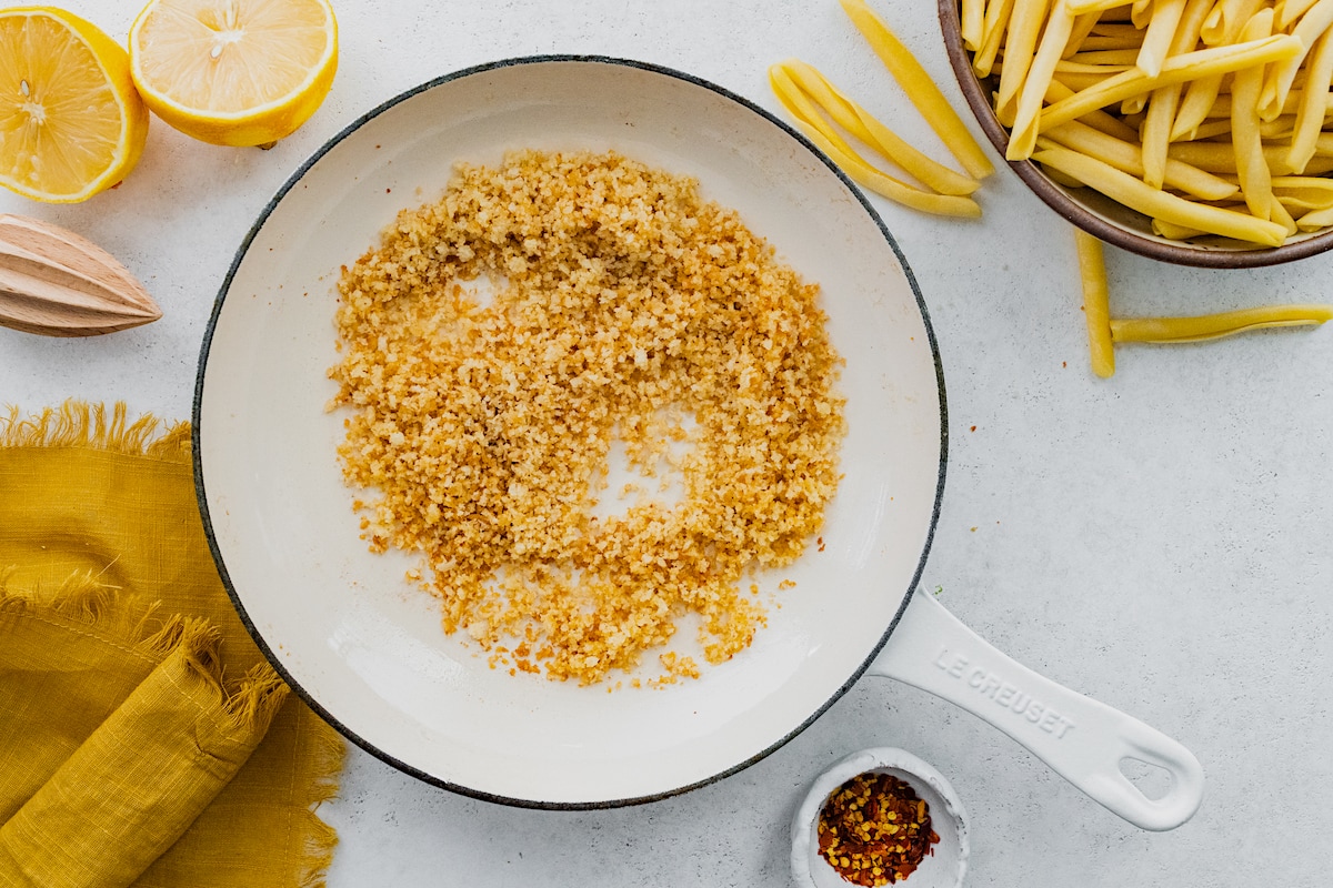 toasted breadcrumbs in a white skillet with pasta in a bowl and lemons.