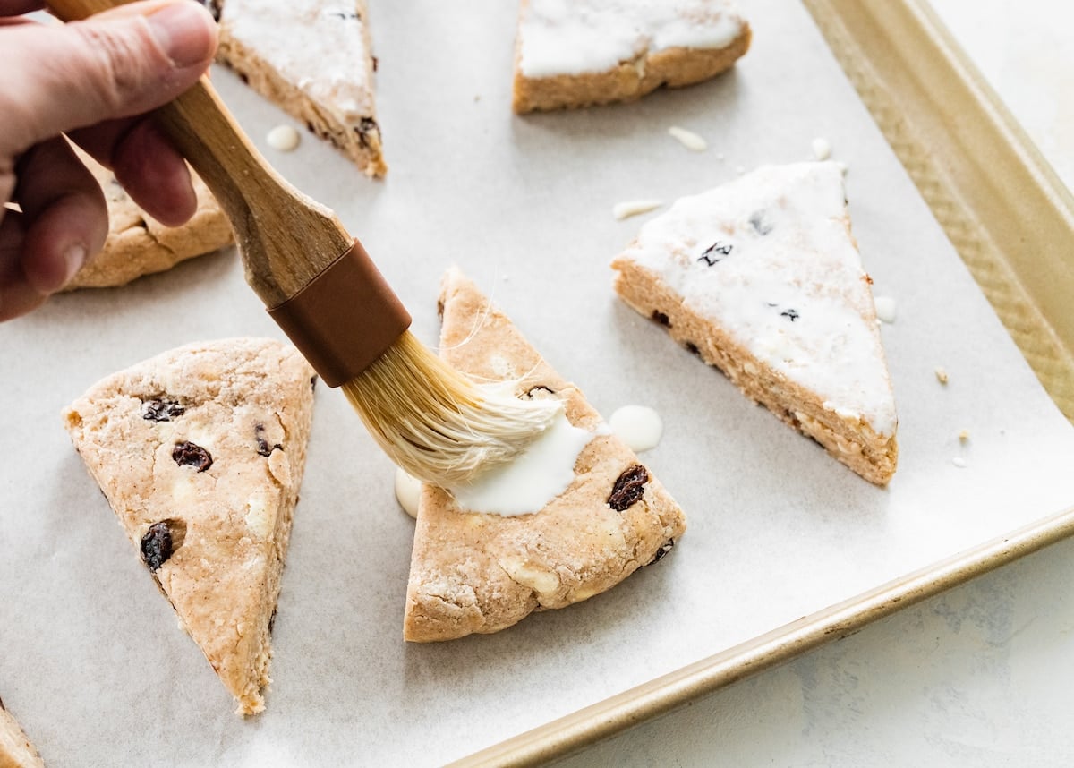 unbaked cinnamon raisin scones on baking sheet being brushed with heavy cream.