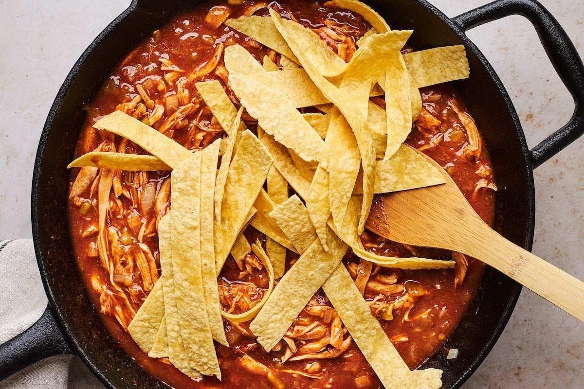 tortilla strips being stirred into skillet chicken enchiladas with wooden spoon.