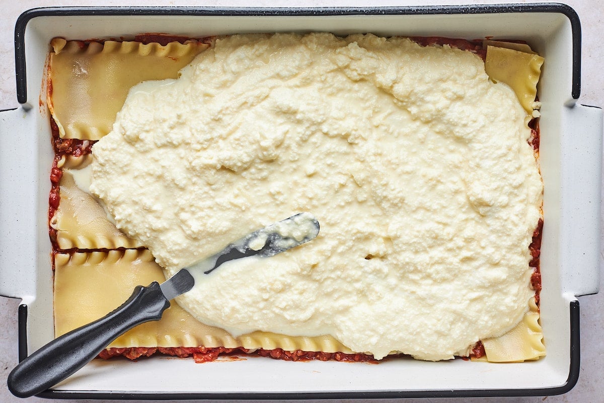 topping cooked lasagna noodles with cheesy ricotta mixture in baking pan.