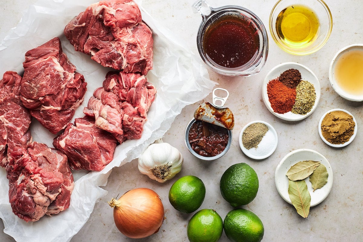 Barbacoa ingredients including beef chuck roast, onion, garlic, limes, beef broth, chipotles in adobo sauce, and spices.