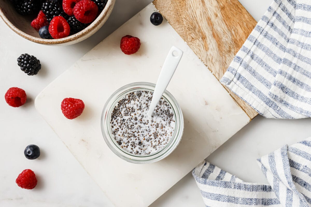 chia pudding being stirred in small glass jar on marble board with tea towel and berries.
