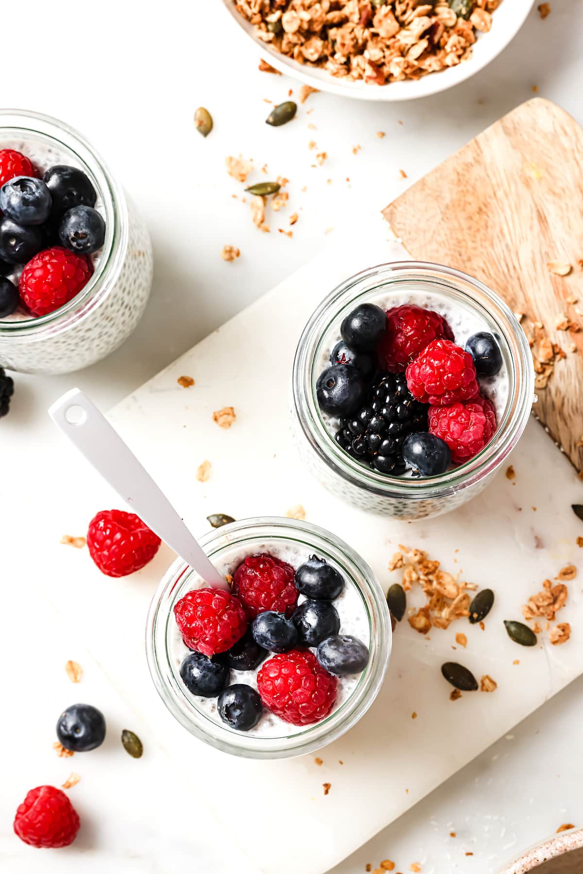 chia pudding in 3 small glass jars with berries and granola on marble board.