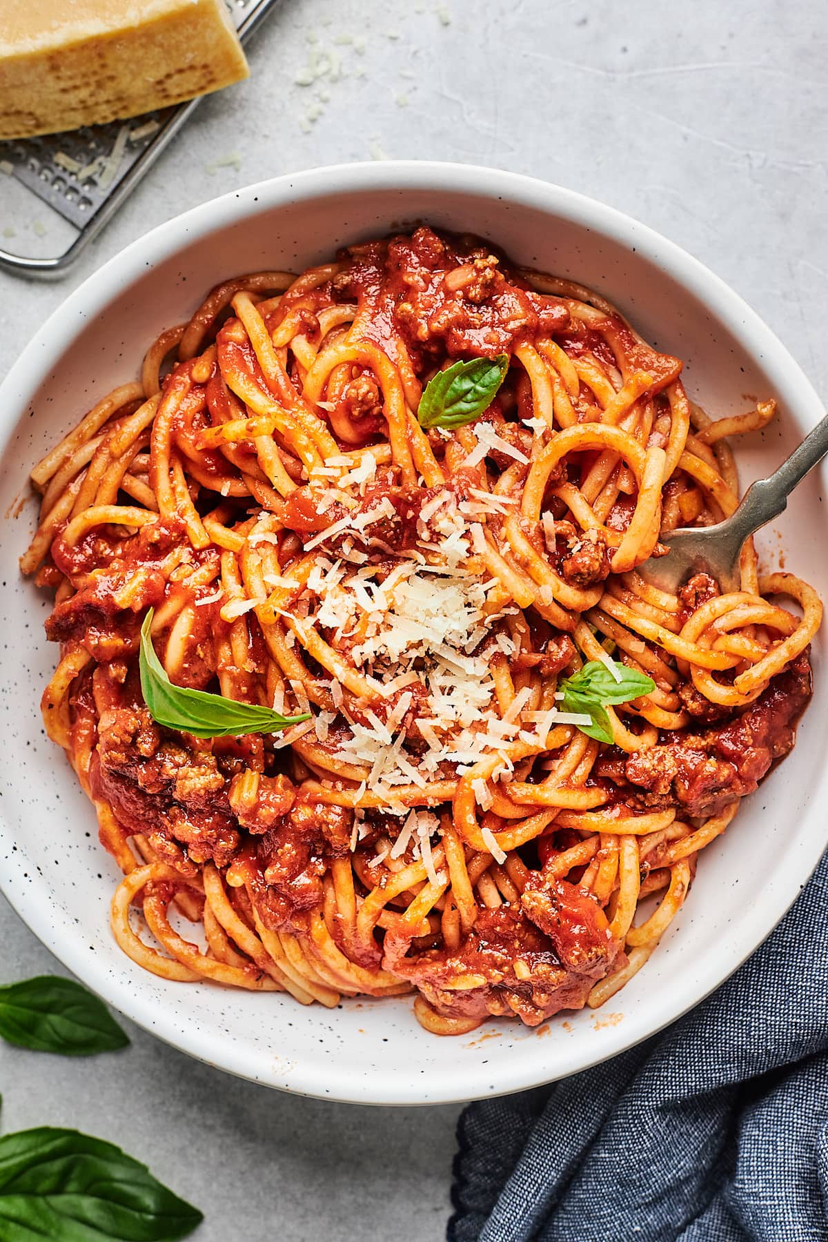 spaghetti with meat sauce topped with Parmesan cheese and basil in white bowl with fork.
