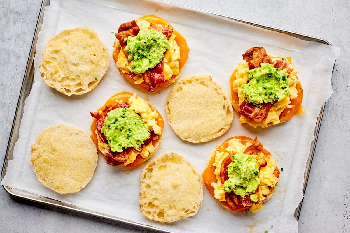 breakfast sandwiches on baking sheet with eggs, cheese, bacon, and mashed avocado.