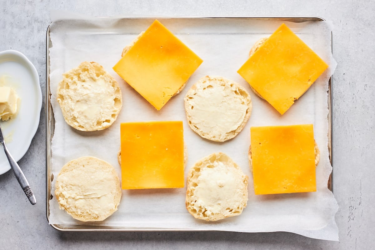 buttered English muffins on baking sheet with cheese slices to make breakfast sandwiches.
