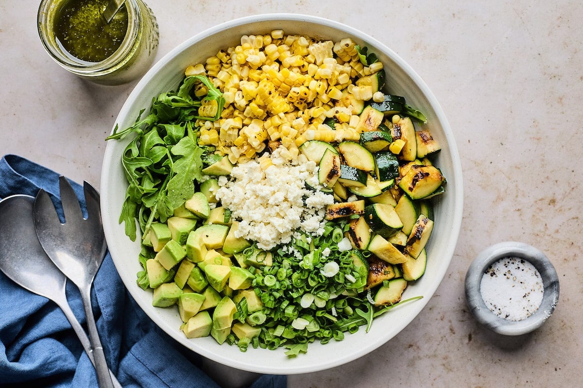 grilled corn, zucchini, green onion, avocado, arugula, and queso fresco in large white bowl.