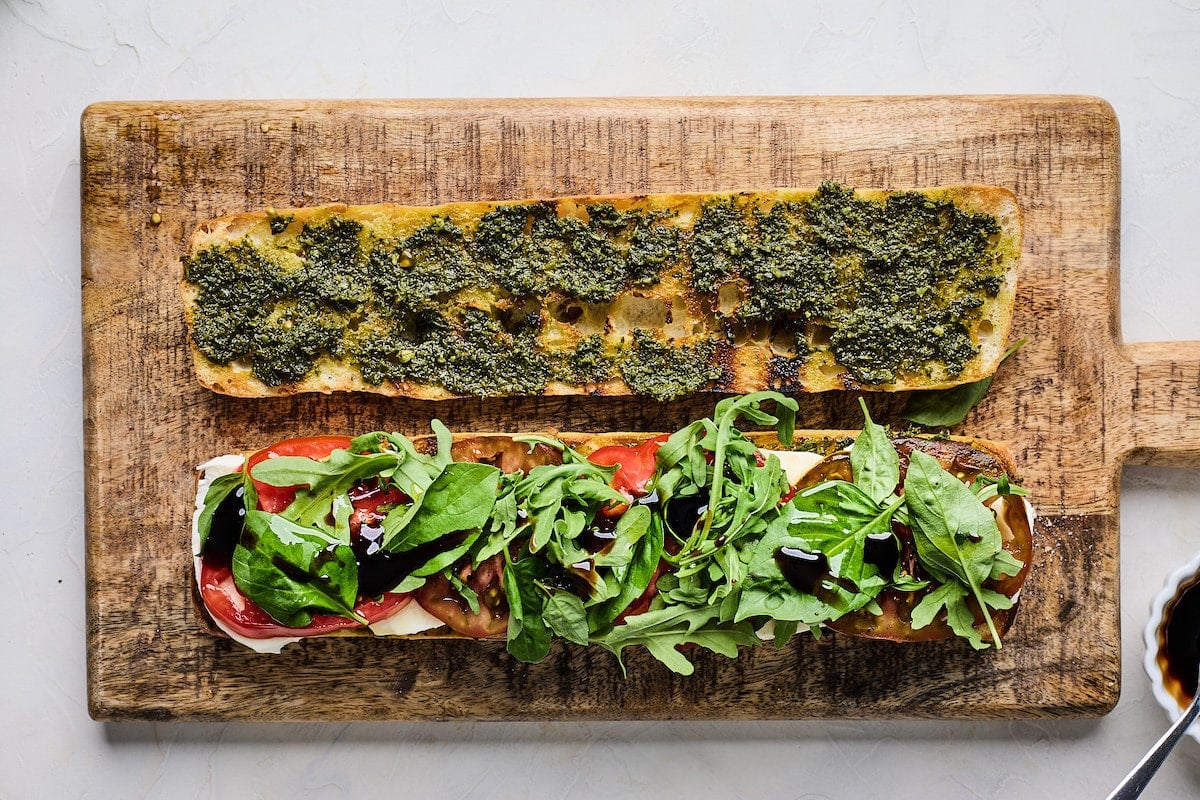 ciabatta bread topped with pesto, fresh mozzarella, tomatoes, basil, arugula, and balsamic glaze on wood board.