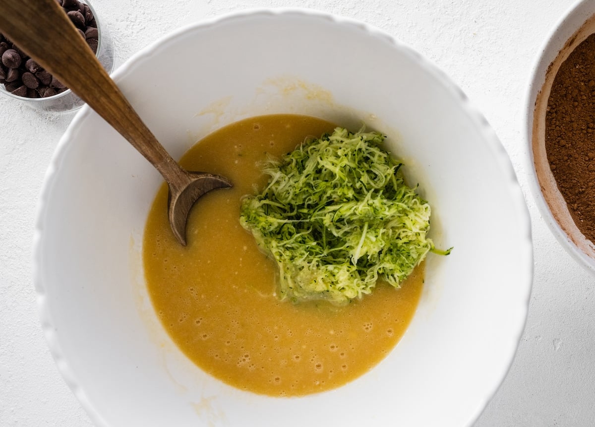 liquid ingredients and shredded zucchini in mixing bowl with wooden spoon.