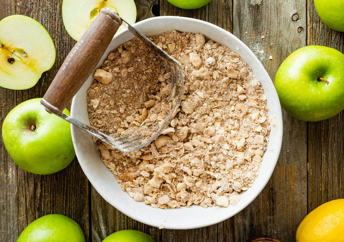 apple crisp topping in bowl with pastry cutter mixing in butter with green apples.