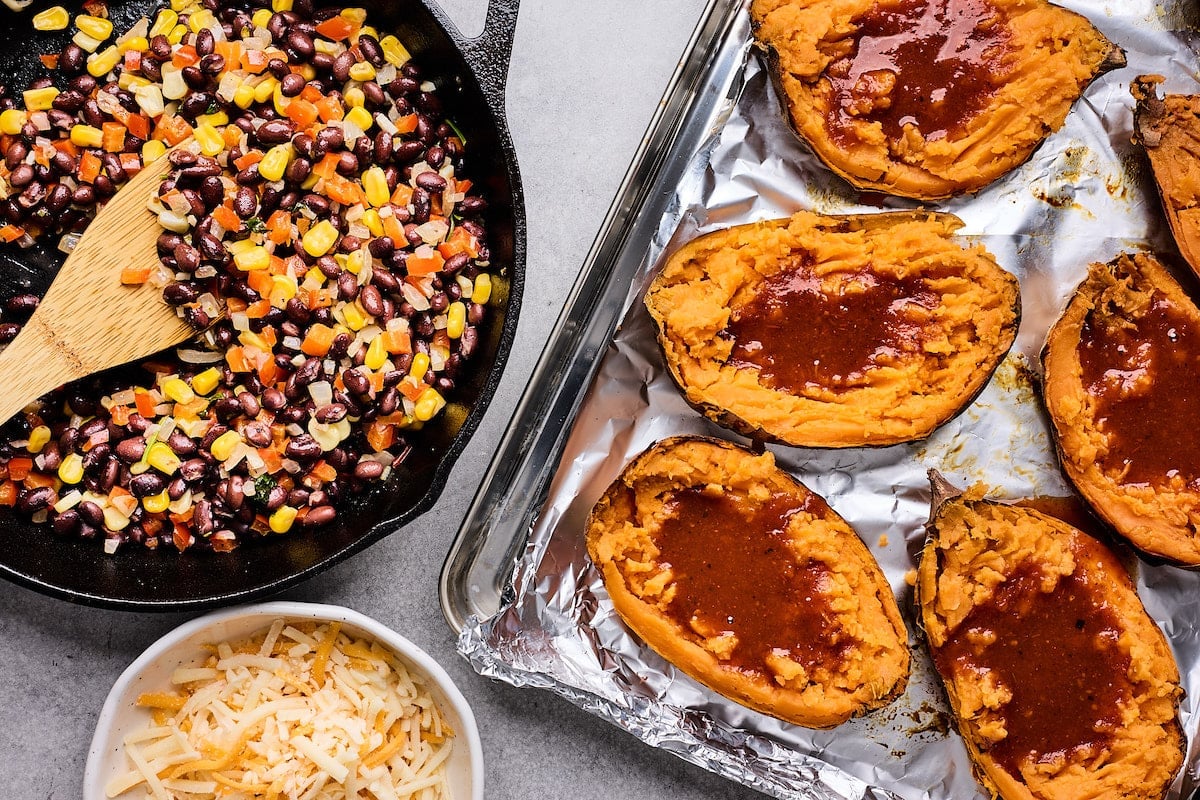 baked sweet potatoes on baking sheet with enchilada sauce and black bean filling in cast iron skillet.