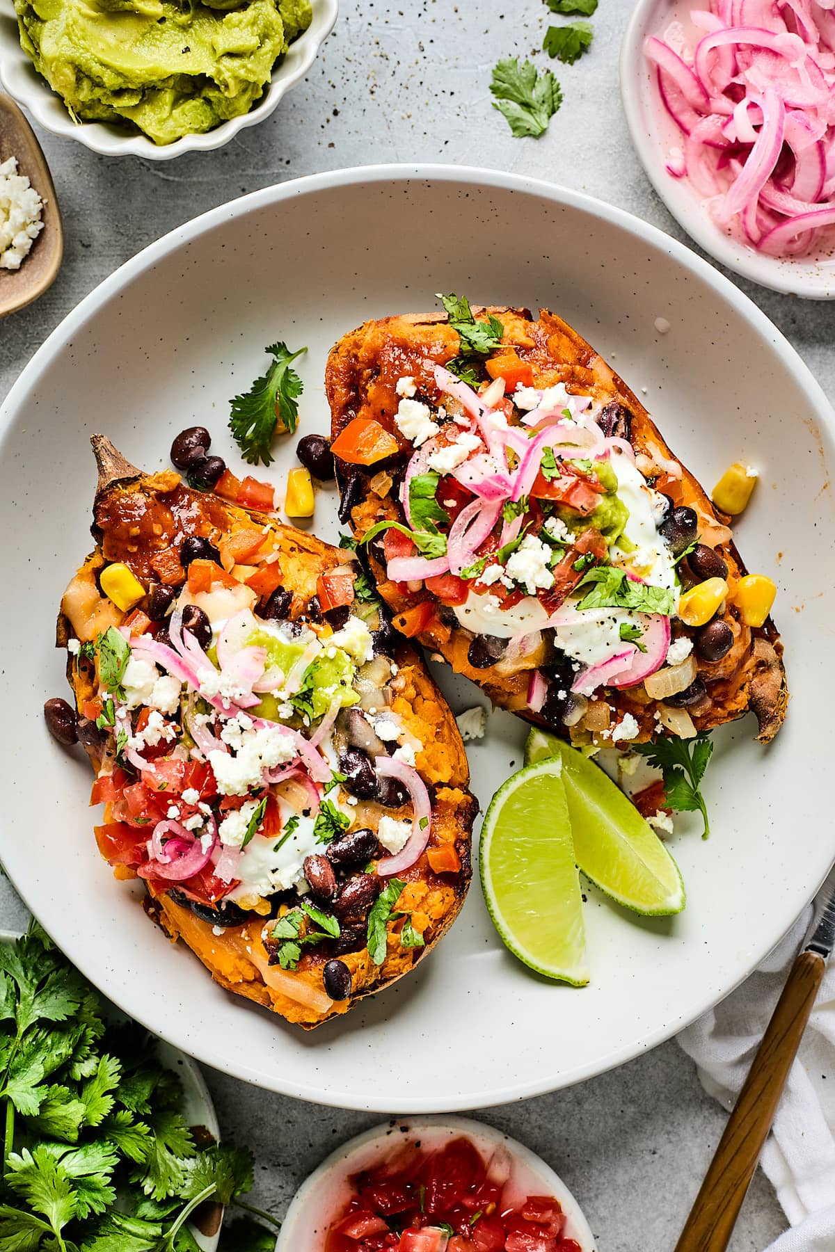 stuffed sweet potatoes on plate with black beans, cheese, tomatoes, pickled red onions, corn, cilantro, and lime wedges.