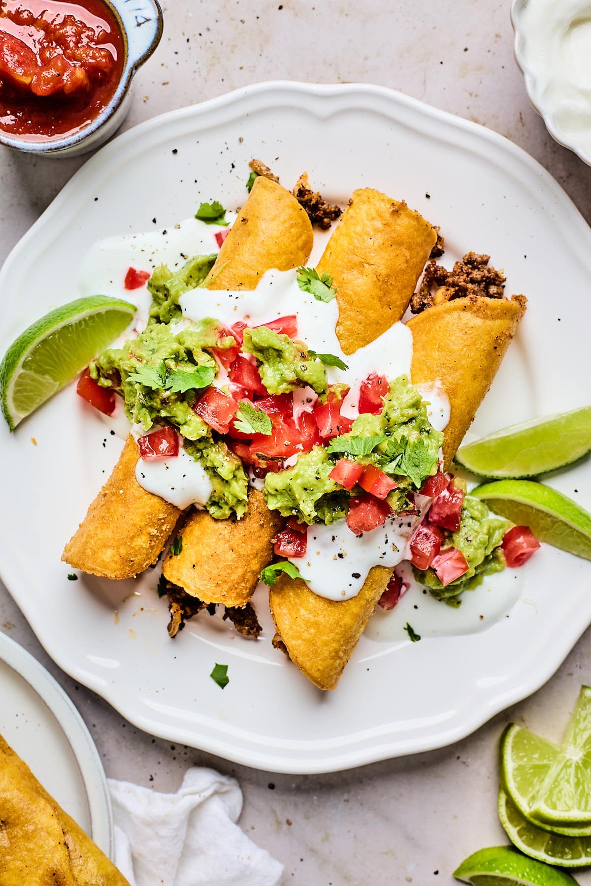 beef taquitos on plate with sour cream, tomatoes, guacamole, cilantro, and lime wedges.