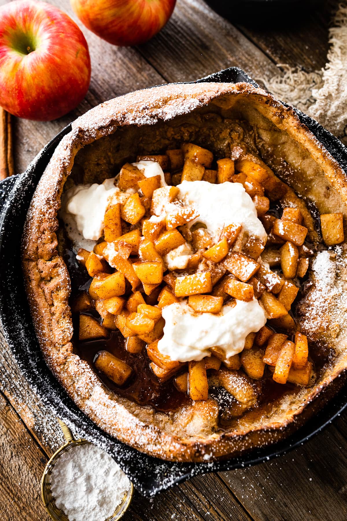 brown butter dutch baby pancake in cast iron skillet with cinnamon apples, whipped cream, and confectioner's sugar.